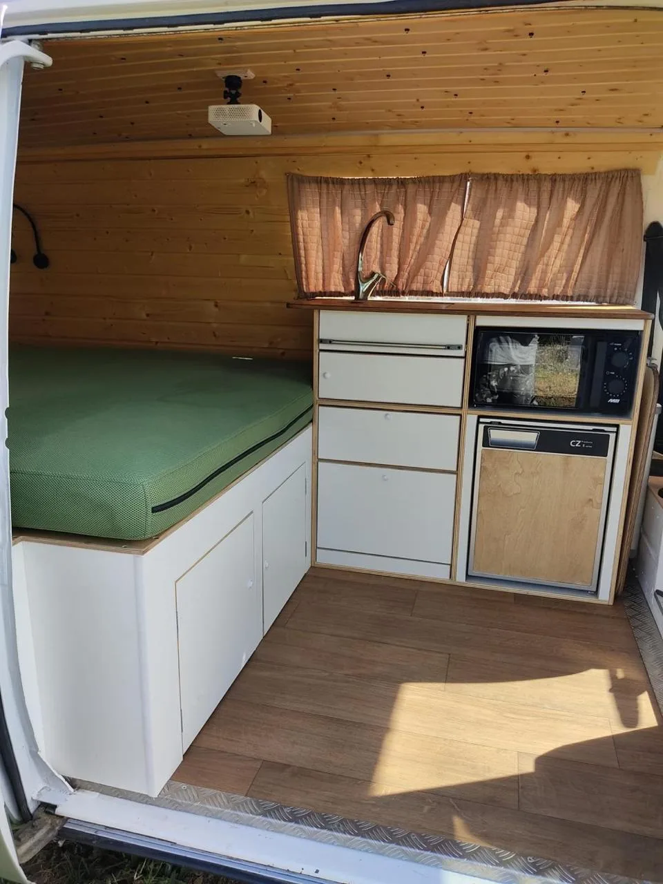 Side-door interior view with bed, kitchen and wooden ceiling